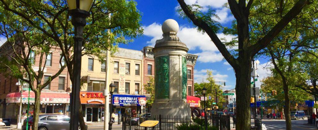 A quaint urban street scene featuring a central stone monument with a spherical top. Surrounding it are various storefronts, including a hardware store and other local businesses. The sky is blue with scattered clouds, and trees frame the view.