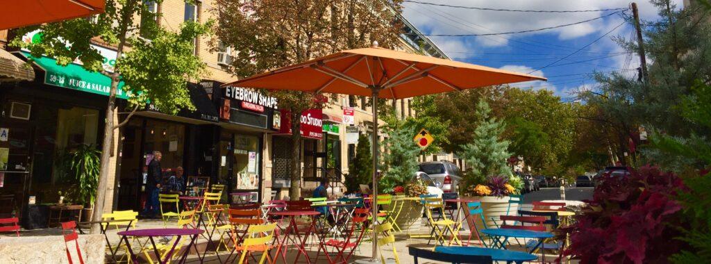 Outdoor café with colorful mismatched chairs and tables under large orange umbrellas lining a quaint street. Shopfronts, a pharmacy, and a convenience store are visible, with greenery and trees in the background under a clear blue sky.